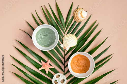 Cosmetic clay masks on peach color background with tropical palm leaf. SPA natural organic beauty products, summer facial skin care concept. Top view, flat lay.