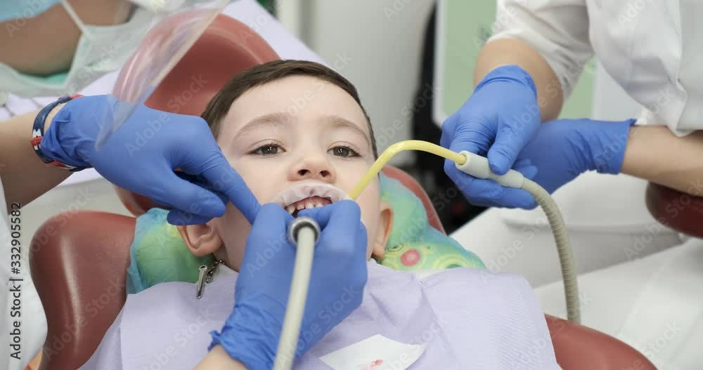 Boy of preschool age at udantist's appointment, doctor and his assistant treat teeth to patient, brushing, hygiene of oral cavity, medical care, dental drill and suction, open mouth, rubber gloves Boy of preschool age at udantist's appointment, doctor and his assistant treat teeth to patient, brushing, hygiene of oral cavity, medical care, dental drill and suction, open mouth, rubber gloves