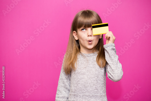Joyful little blonde shy face girl 6-7 years old hold credit bank card isolated on pink. Childhood lifestyle concept.