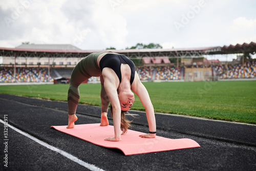 Young brunette sportswoman, wearing black top and green leggings, standing in gymnastic bridge on city stadium on summer morning, Stretching training outside on pink yoga mat. Healthy life concept © Natalia