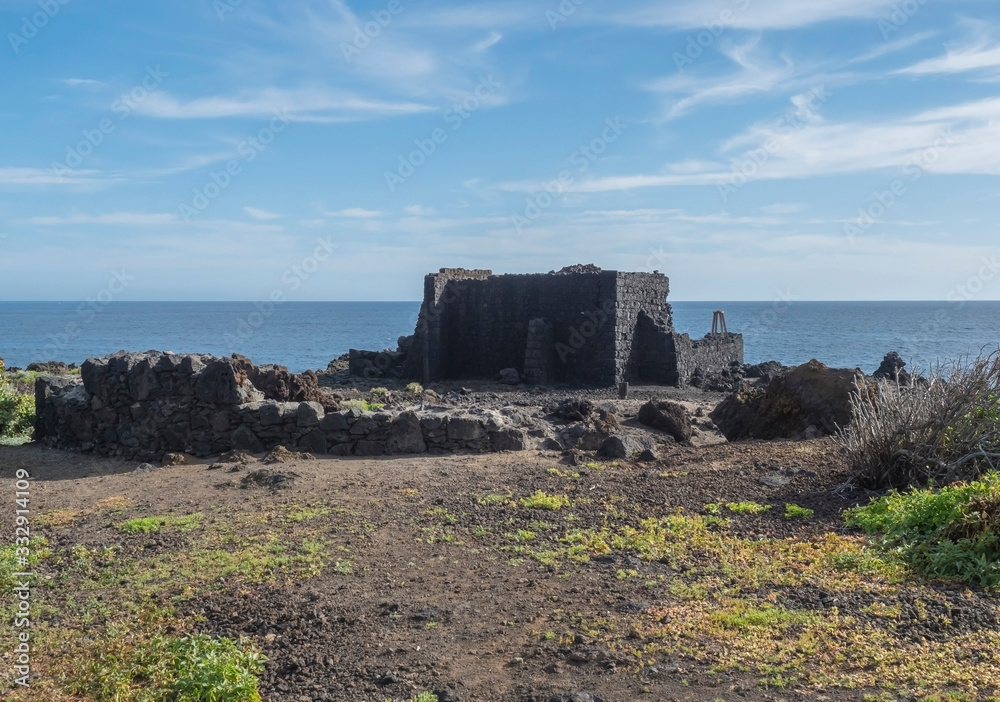 Old remains of ruined fortification from lava stone on sea shore with blue sky at Canary islands village.
