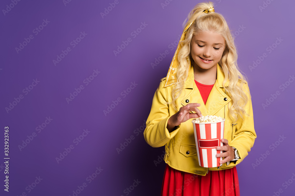 Fototapeta premium smiling kid holding bucket with popcorn on purple background