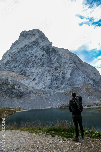 A man in hiking outfit standing by the Wolayer Lake in Austrian Alps. There is massive, rocky mountain on the other side of the lake. New day beginning. Soft reflections in the lake. Happiness