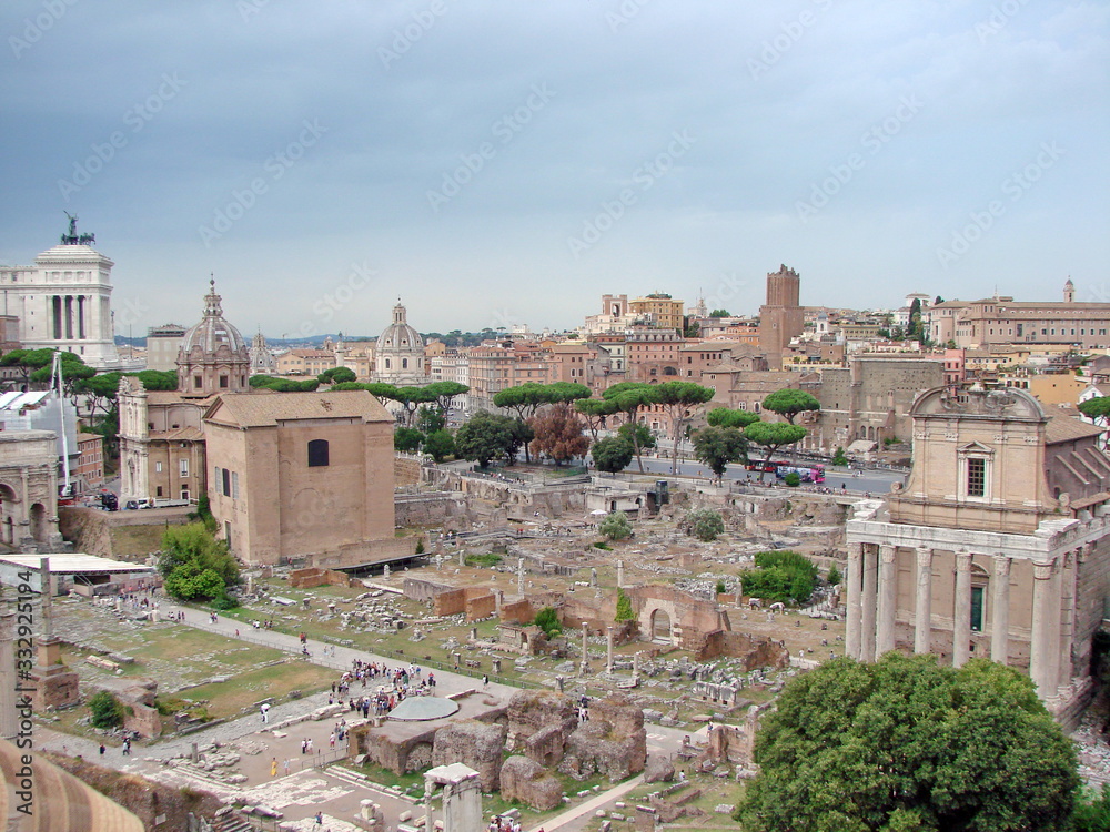 Landscape of millennial columns and dilapidated majestic buildings of mighty Rome in the past, against the backdrop of modern areas under a cloudy sky on the horizon.