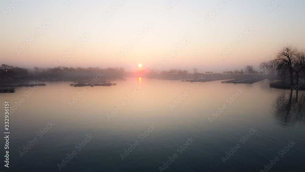 Wide angle shot of calm water in lake inlet with empty boat docks as sun rises over fog covered trees in the early morning in Chicago Illinois