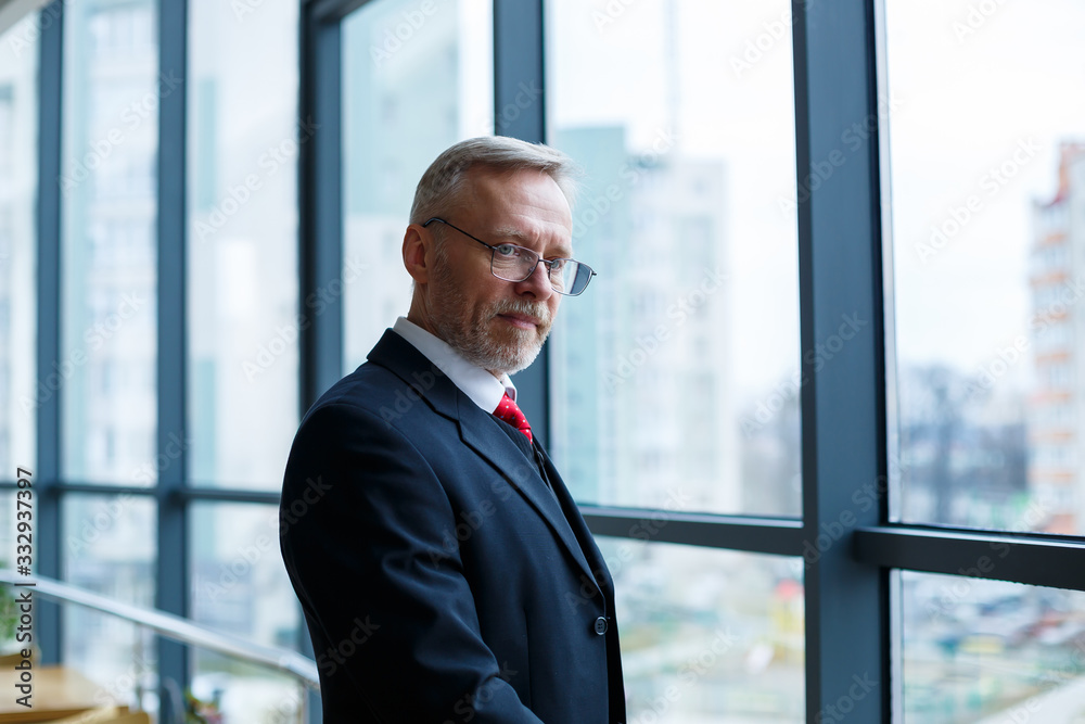 Fototapeta premium Adult male businessman, teacher, mentor looks out the large window of his office. Work in the office. Development of a new business project
