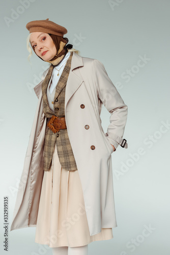 Elegant senior woman with hand in pocket of trench coat looking at camera isolated on grey