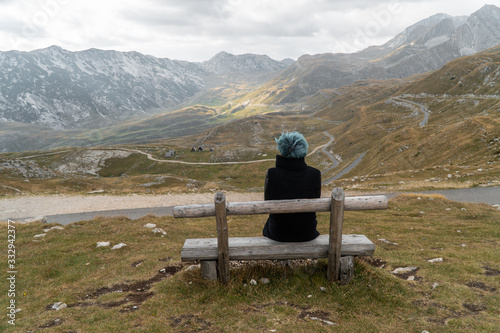 Young woman with blue hair in a black coat sits on a wooden bench by the road in the mountains