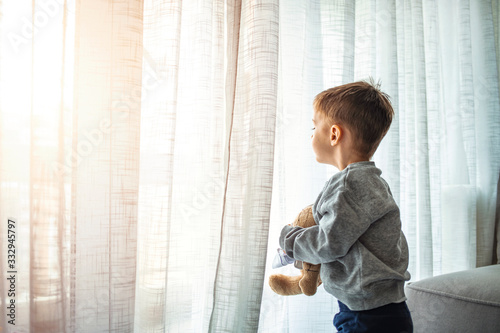 Quarantine, threat of coronavirus, virus protection, pandemic. Child and his teddy bear both in protective masks sits on windowsill inside house and looks out the window. Coronavirus epidemic.