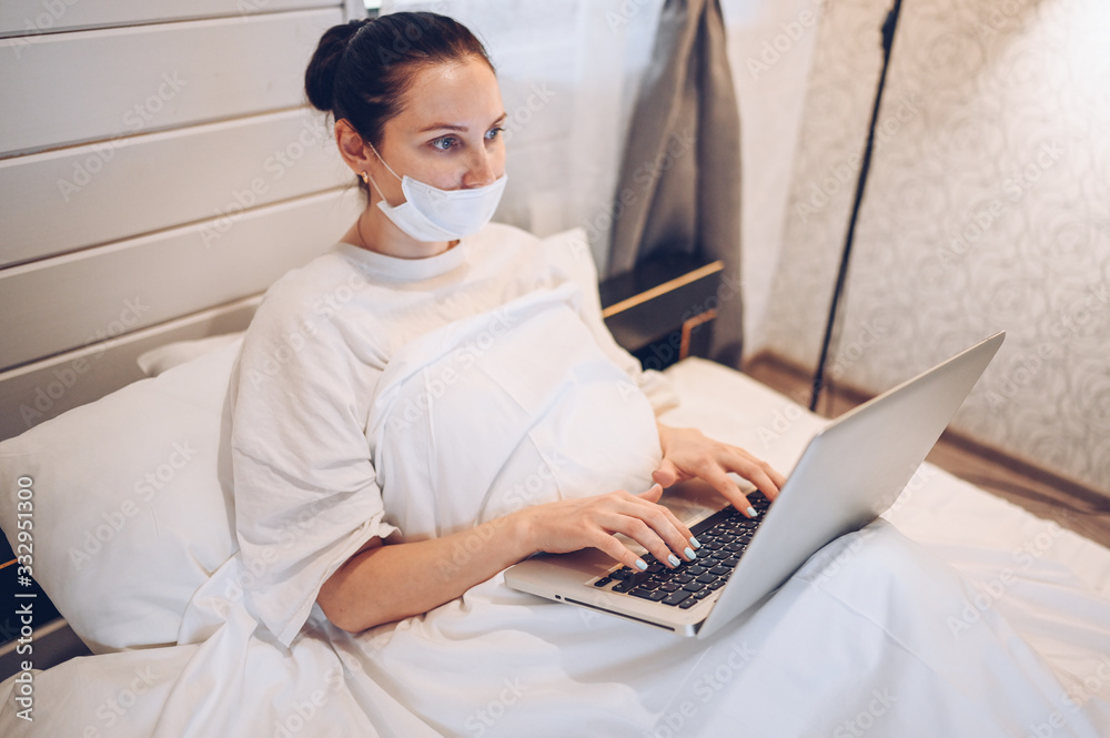 Young european woman in face medicine mask working on a laptop in bedroom during coronavirus isolation home quarantine. Covid-19 pandemic Corona virus. Distance online work from home concept. Young european woman in face medicine mask working on a laptop in bedroom during coronavirus isolation home quarantine. Covid-19 pandemic Corona virus. Distance online work from home concept.