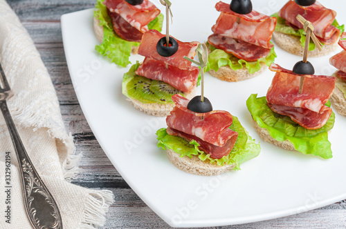 Canapes with carbonade, kiwi and lettuce on a white wheat bread. Garnished with Olive. On a white plate. Near a fork and a napkin.