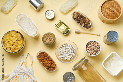 Creative flatlay with pantry staples. Glass jars with pasta, beans and chickpeas, canned goods, nuts and dried mushrooms in reusable containers. Top view pattern with basic products