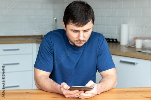 Handsome bearded man in a blue T-shirt sits behind a wooden table and reads an electronic book against a blurred background of a modern kitchen