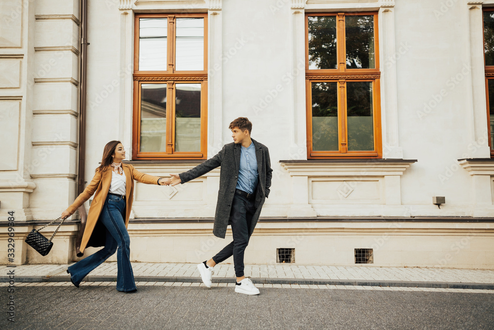 Happy to be together. Side view of beautiful young couple holding hands and looking at each other with smile while walking through the city street. Couple on the go. Follow me. Stylish couple.