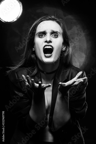 Beautiful braless girl with red lips, wearing a black blazer, screams, stretching forward her hands on a dark background, posing next to a light lamp. Advertising, trendy, monochrome design.
