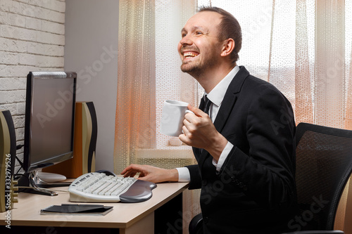 Young male manager works at a computer in the office. Holds a glass of tea in his hand. Chatting with friends on social networks and laughing. Telework. A man in a black jacket and white shirt.