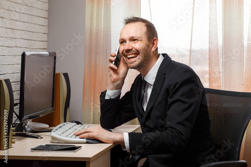 Young male manager works at a computer in the office. Talking on a cell phone with a client, discussing a business project and laughing. Telework. A man in a black jacket and white shirt. 