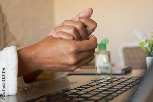 Hands of Brazilian black woman cleaning hands with sanitizer gel in front of laptop at home. Sleeves of robe. Working from home due Coronavirus or Covid-19 quarantine.