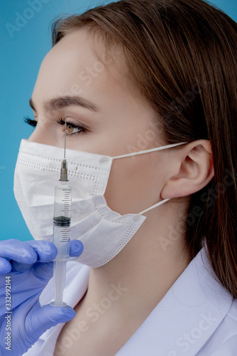 Woman doctor in a white medical coat  mask  hat and gloves holds a syringe with medicine in her hands. Standing on a white background