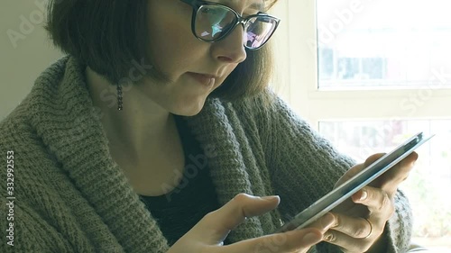 Young caucasian woman in cardigan and casual clothes wearing black frame eyeglasses and using tablet at home