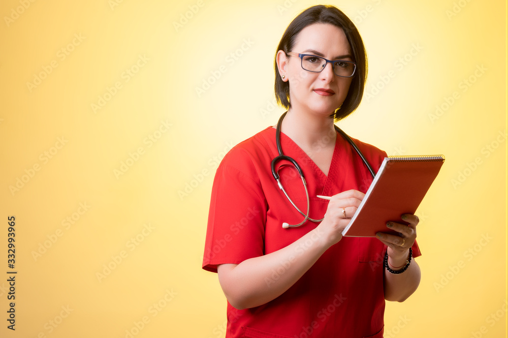 Beautiful woman doctor with stethoscope, wearing red scrubs takes notes, looking at the camera Beautiful woman doctor with stethoscope, wearing red scrubs takes notes, looking at the camera