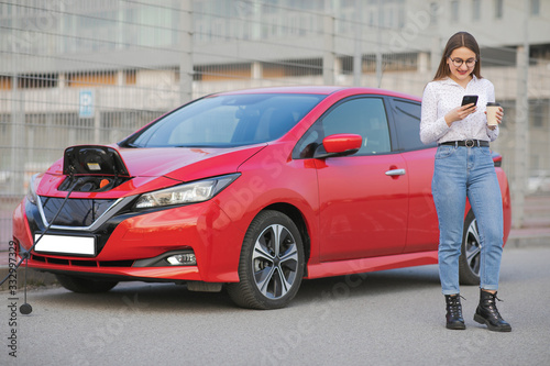 Electric car charging in street. Ecological Car Connected and Charging Batteries. Girl Use Coffee Drink While Using SmartPhone and Waiting Power Supply Connect to Electric Vehicles for Charging