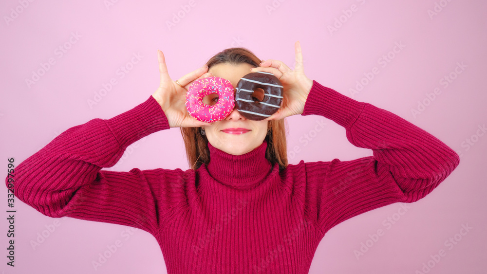 Adult female in read sweater smiling and keeping sweet round donuts near eyes looking at camera against pink background Adult female in read sweater smiling and keeping sweet round donuts near eyes looking at camera against pink background