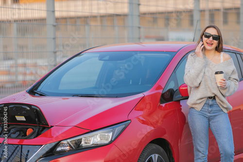 Stylish girl stands with phone near her red electric car and waits when vehicle will charged. Connecting the Charger Plug of an Electric Car.