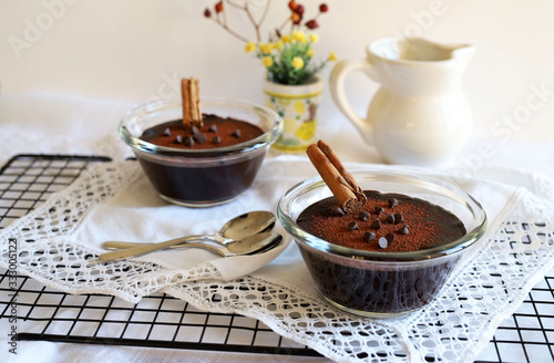 Primo piano di budino al cioccolato fatto in casa cosparso di cacao in polvere, decorato con gocce di cioccolato e stecca di cannella.