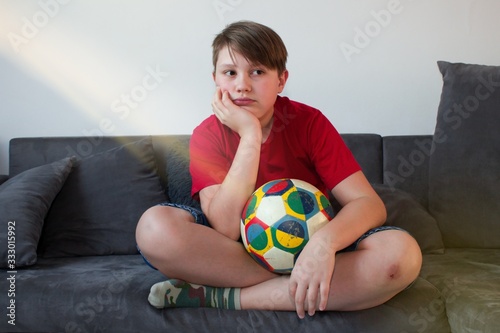 Bored child sitting on the couch and holding a football ball.Stay at home. Teenage boy bored sitting with kicking ball on couch. Quarantine due to coronavirus pandemic.