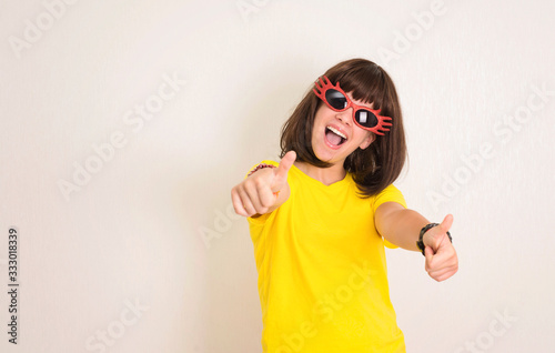 Comic, charming, positive, girl showing thumbs up gesture. Crasy teen girl wearing party glasses. Playful teenager girl with funny large glasses. Party time.