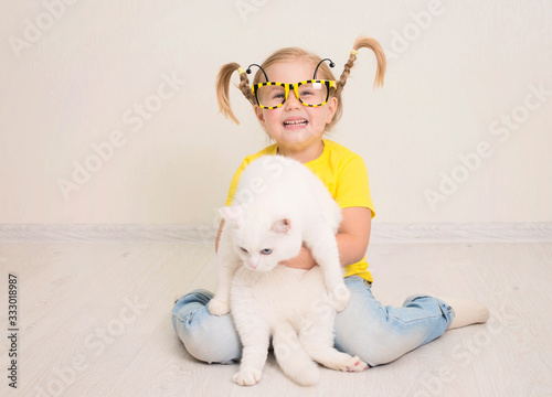 Little girl in funny bee glasses and yellow t-shirt and with braids smiling happy hugging her cat. photo