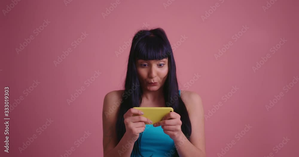 Positive smiling young brunette woman in blue shirt playing games on mobile phone isolated on pink background Positive smiling young brunette woman in blue shirt playing games on mobile phone isolated on pink background