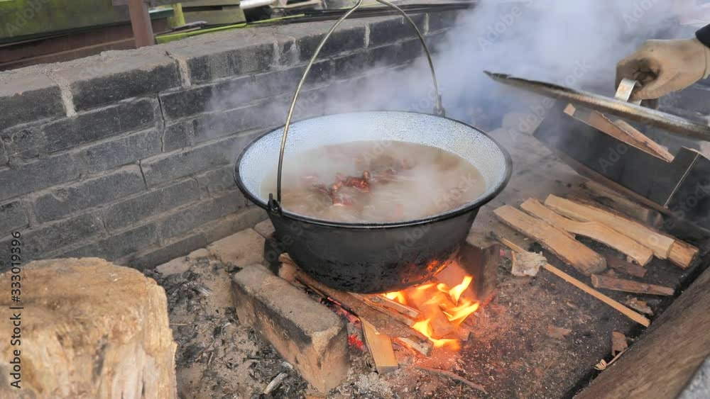 Round view onto keetle above BBQ fire. Big enamel pot with chicken broth hanging on the yard.