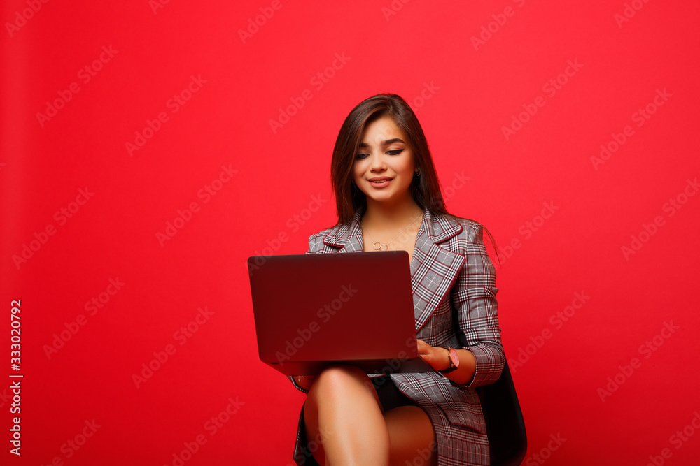 Smiling young brunette woman girl in modern suit posing isolated on red background. Holding and working on laptop. Studio portrait. People lifestyle concept. Mock up copy space. Smiling young brunette woman girl in modern suit posing isolated on red background. Holding and working on laptop. Studio portrait. People lifestyle concept. Mock up copy space.