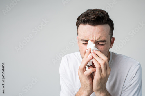 Young man isolated over background. Portrait of guy sneezing into white napkin. Sickness and coronavirus danger. Ill guy on picture. Running nose and bad feeling.
