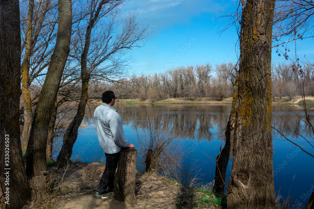 A middle-aged man stands with his back and looks at the spring river in the forest. Bright sky and spring forest are reflected in the river