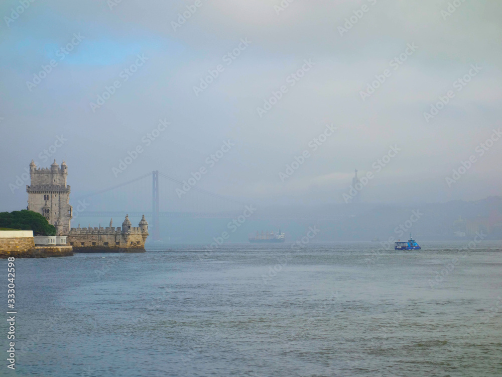 Skyline of Lisbon in the background on the left the tower of Belem, the bridge April 25 crossing the river Tejo, the boats and the Christ the King on a foggy day.