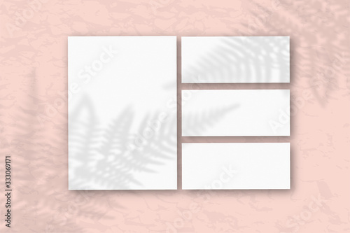 Several horizontal and vertical sheets of white textured paper against a pink wall. Mockup overlay with the plant shadows. Natural light casts shadows from the fern leaves. Flat lay  top view