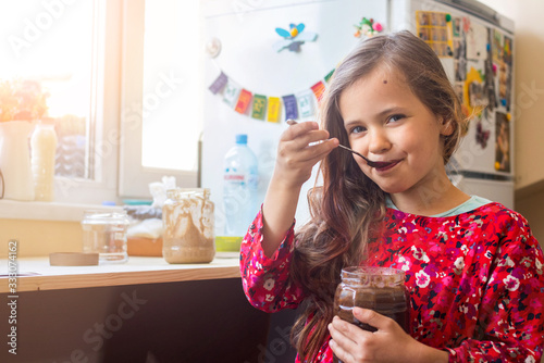 Cute happy young smiling girl eat homemade sweet chocolate paste spread from big glass jar with joy and pleasure
