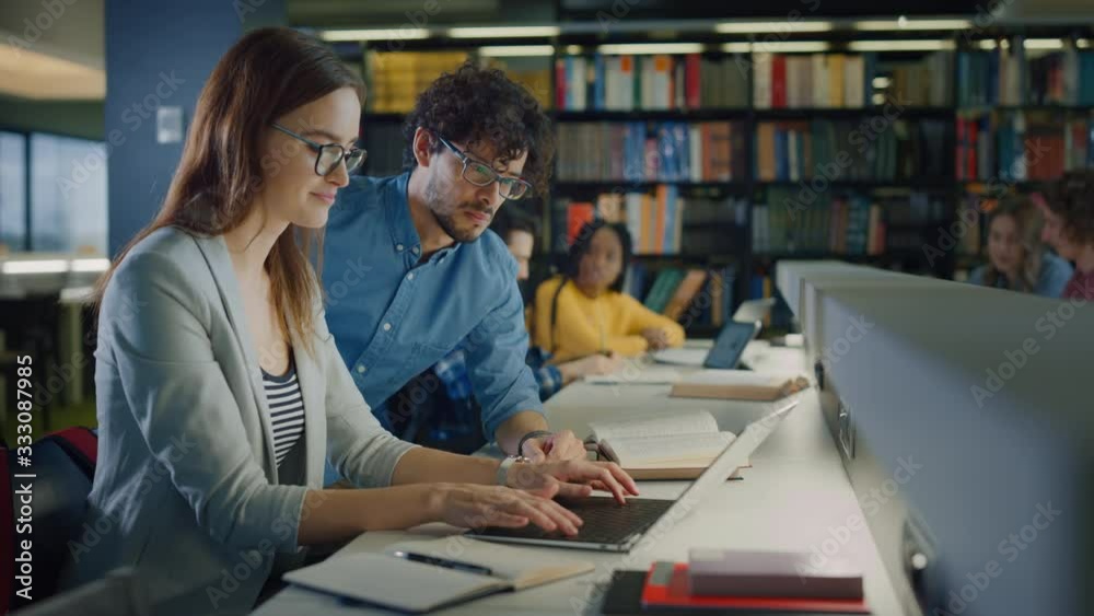 University Library: Talented Caucasian Girl uses Laptop, Smart Helpful Classmate Explains and Advices Her with Class Assignment. Happy Diverse Students Talking, Learning, Studying Together for Exams