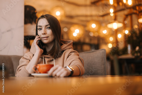 Young woman freelancer in casual clothes talking on the phone at a table in a cozy cafe. Coffee cup on the table. Concept of leisure activity.