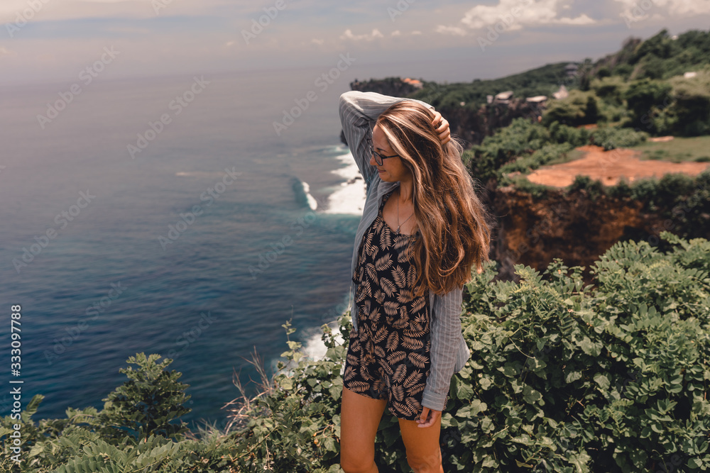 Beautiful girl traveler with long hair and a slender body stands on a cliff of a cliff overlooking the waves of the blue ocean.Bali Island, Indonesia. Travel and tourism concept in exotic and tropical
