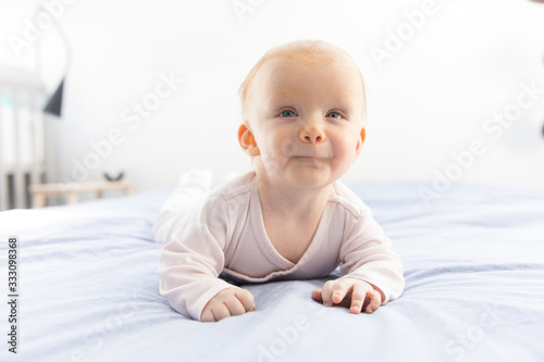 Cheerful adorable red haired baby girl crawling on bedding. Six month child lying on belly in bedroom. Childhood or baby care concept