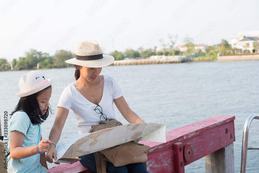 looking away. Outdoor portrait of two girls traveling around new place and searching beautiful sights. wonderful inspired curly woman in hat holding pretty daughter and town by the river map...