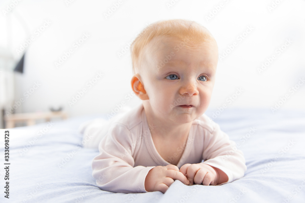 Cute funny red haired baby girl relaxing on bed clothing and looking up. Six month child lying on belly in bedroom. Childhood or baby care concept