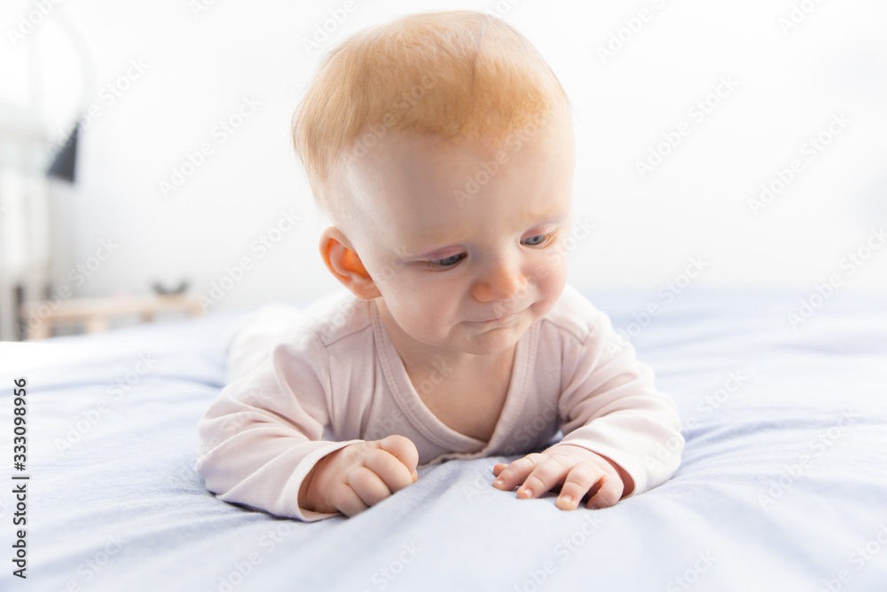 Pensive red haired baby girl crawling on parents bed. Six month child lying on belly in bedroom. Childhood or baby care concept