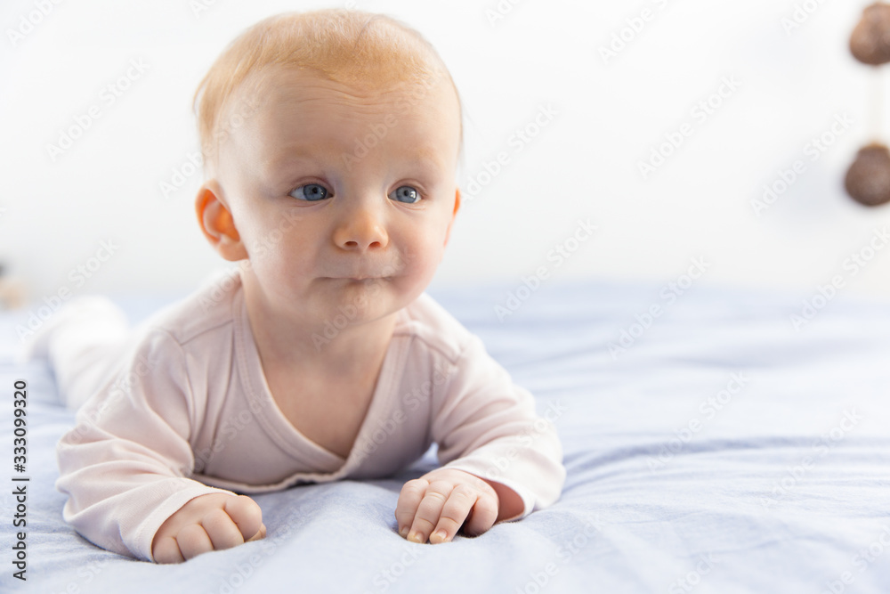 Pensive red haired baby crawling on bedding. Six month child lying on belly at home. Childhood or baby care concept