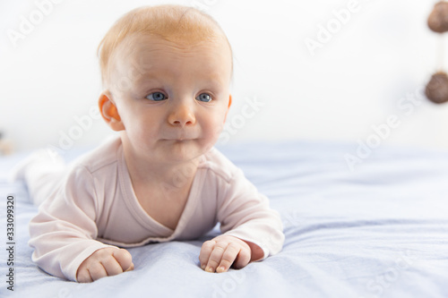 Pensive red haired baby crawling on bedding. Six month child lying on belly at home. Childhood or baby care concept