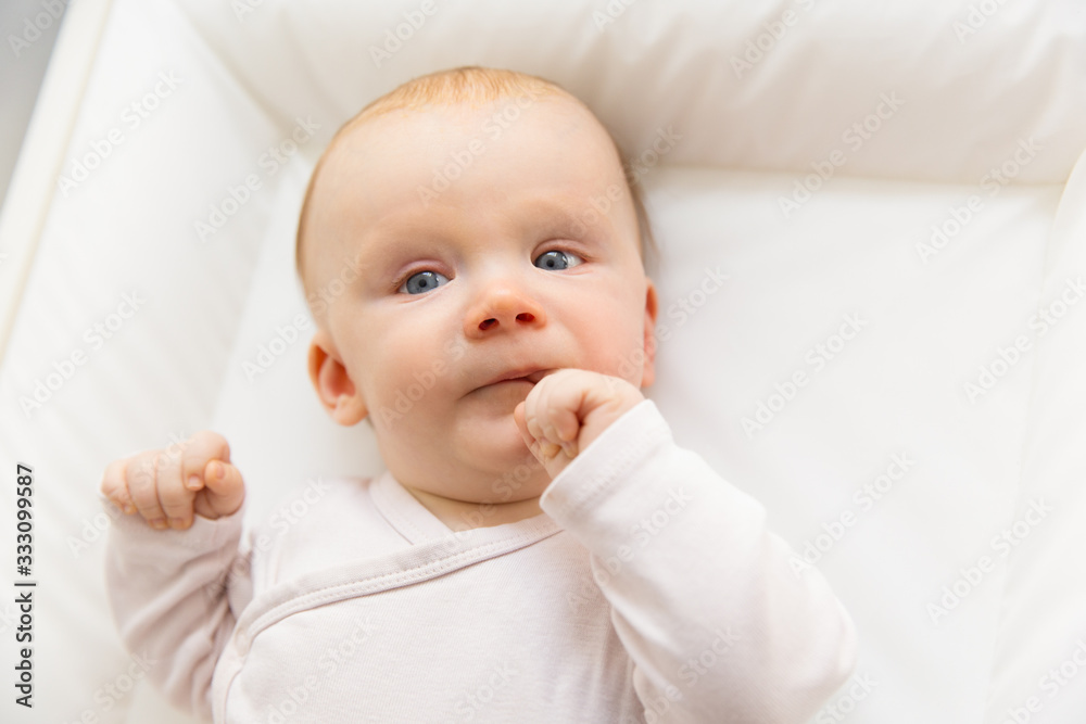 Cute thoughtful red-haired baby holding finger near mouth. Front view of tiny newborn child lying in cradle. Nursery, newborn kid and infancy concept